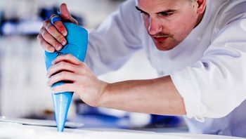 Matlagningskurs - En kock i vit uniform fyller noggrant en form med en blå spritspåse på ett ljust köksbänk, fokuserad på att skapa en detaljerad maträtt.