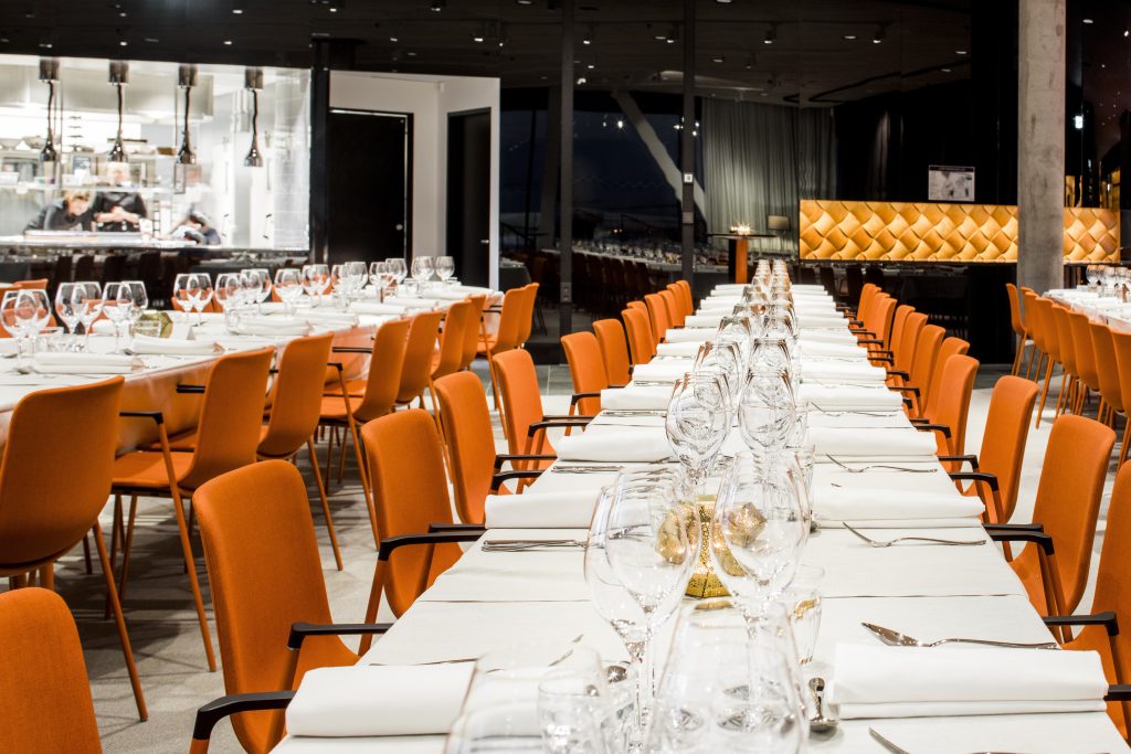 Ett långt matbord dukat med elegant glas och vita tallrikar i en modern restaurang med orange stolar, som visar en sofistikerad och inbjudande atmosfär.