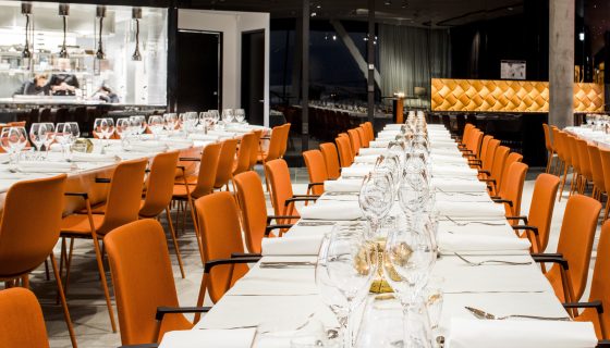 Ett långt matbord dukat med elegant glas och vita tallrikar i en modern restaurang med orange stolar, som visar en sofistikerad och inbjudande atmosfär.